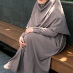 A woman seated outdoors in a gray hijab and abaya, exuding peaceful confidence.